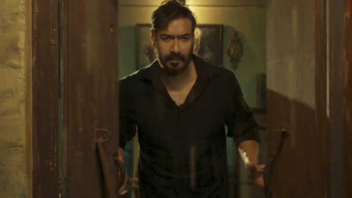 Ajay Devgn in a tense scene from Drishyam: The Conclusion, standing against a dimly lit wall, eyes sharp, jaw set.