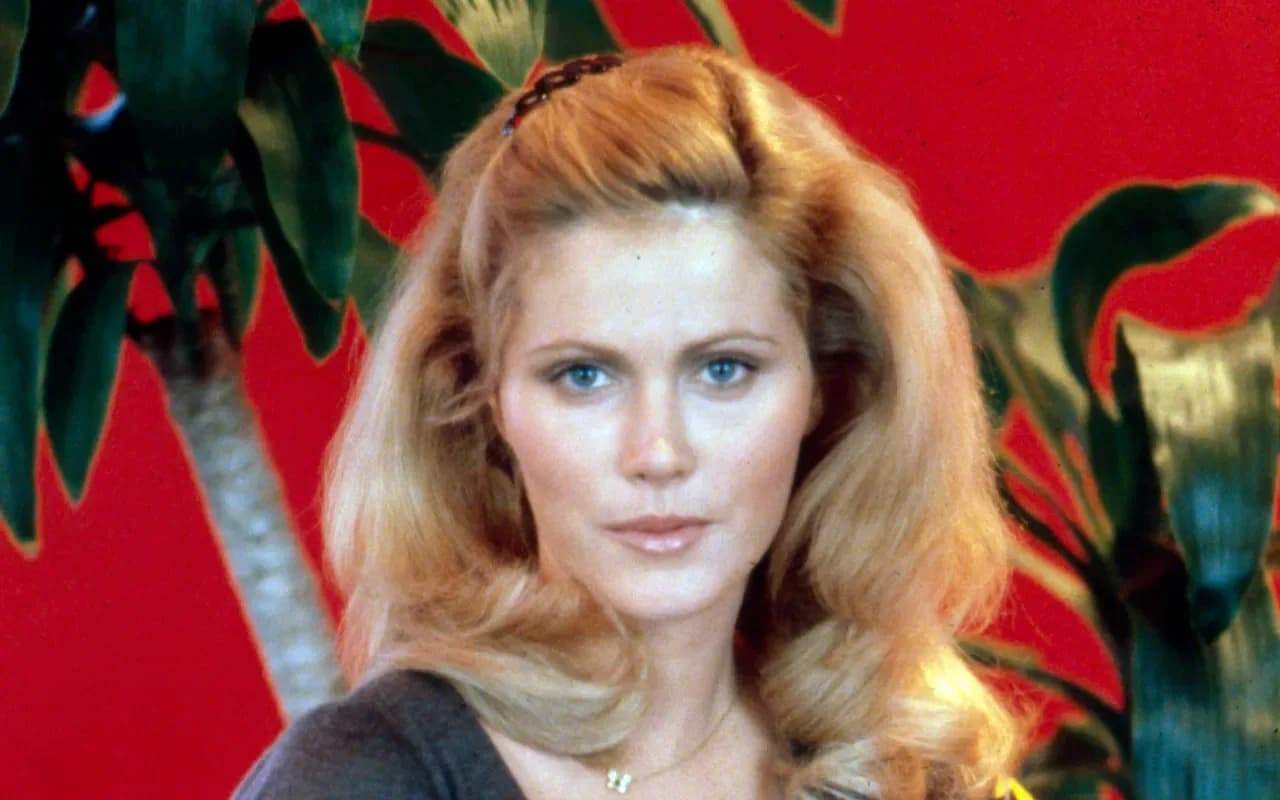Mary Louise Weller in a vintage photo from 'Animal House', standing in front of a sorority house, smiling.