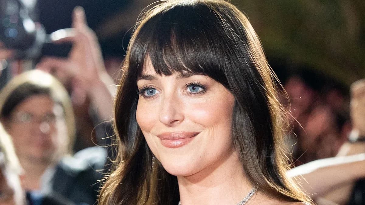 Dakota Johnson in a black dress, standing in front of a neon-lit stage with the SNL logo glowing behind her.