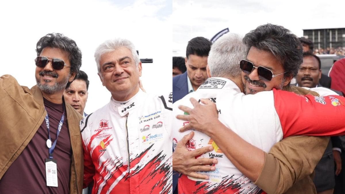 Ajith Kumar in a racing suit embracing Vijay in a brownish jacket at the Silverstone circuit as fans and photographers look on.