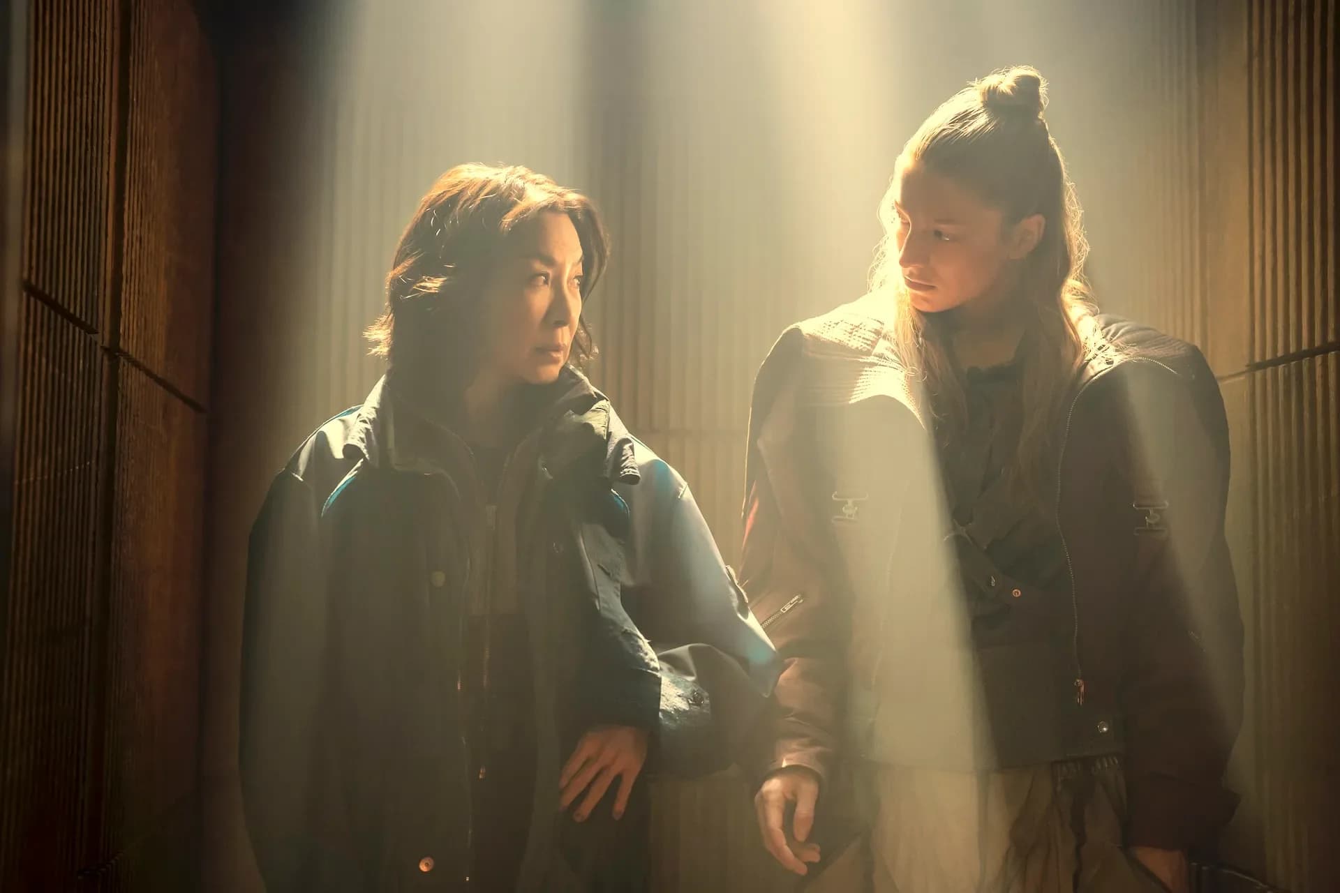 Michelle Yeoh and Hunter Schafer in futuristic streetwear, standing under flickering neon signs in a rain-soaked cityscape.