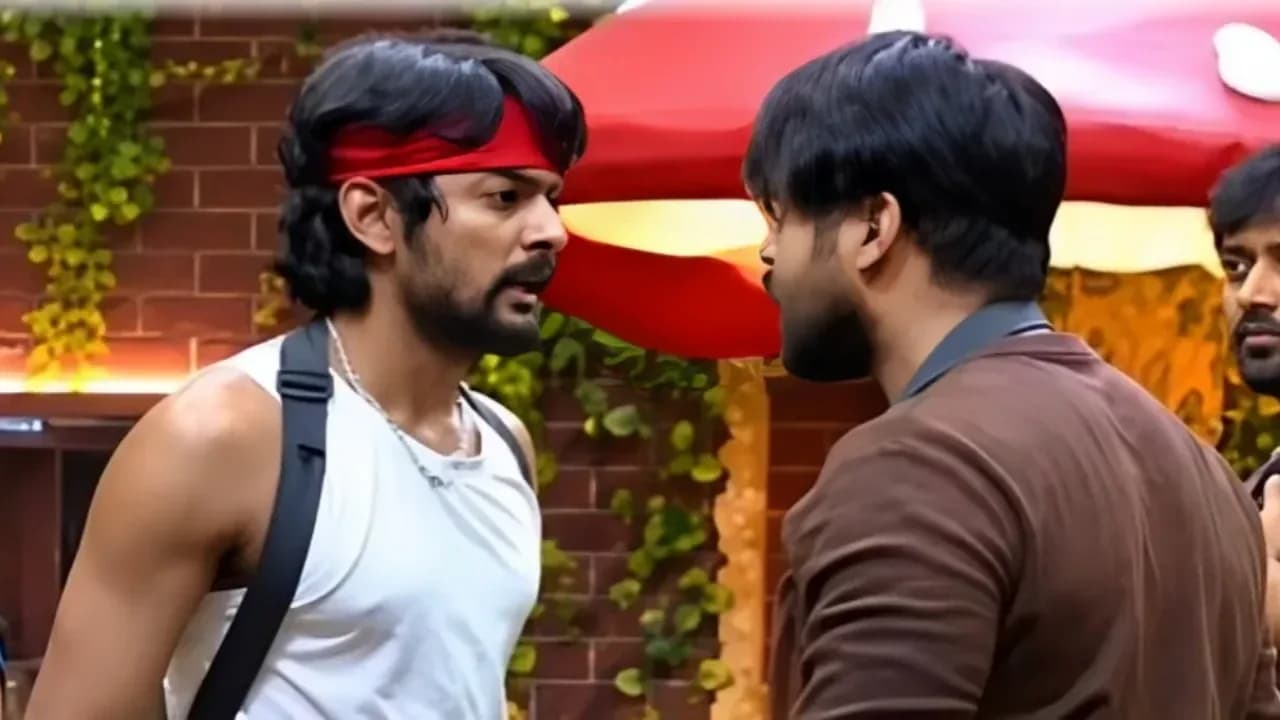 Two men in a heated argument inside a Bigg Boss house, one gesturing aggressively while the other holds his wrist in pain.