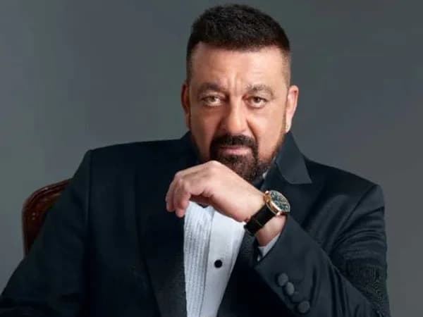 Sanjay Dutt in a black suit, mid-speech on a talk show stage, eyes intense, hands gesturing.