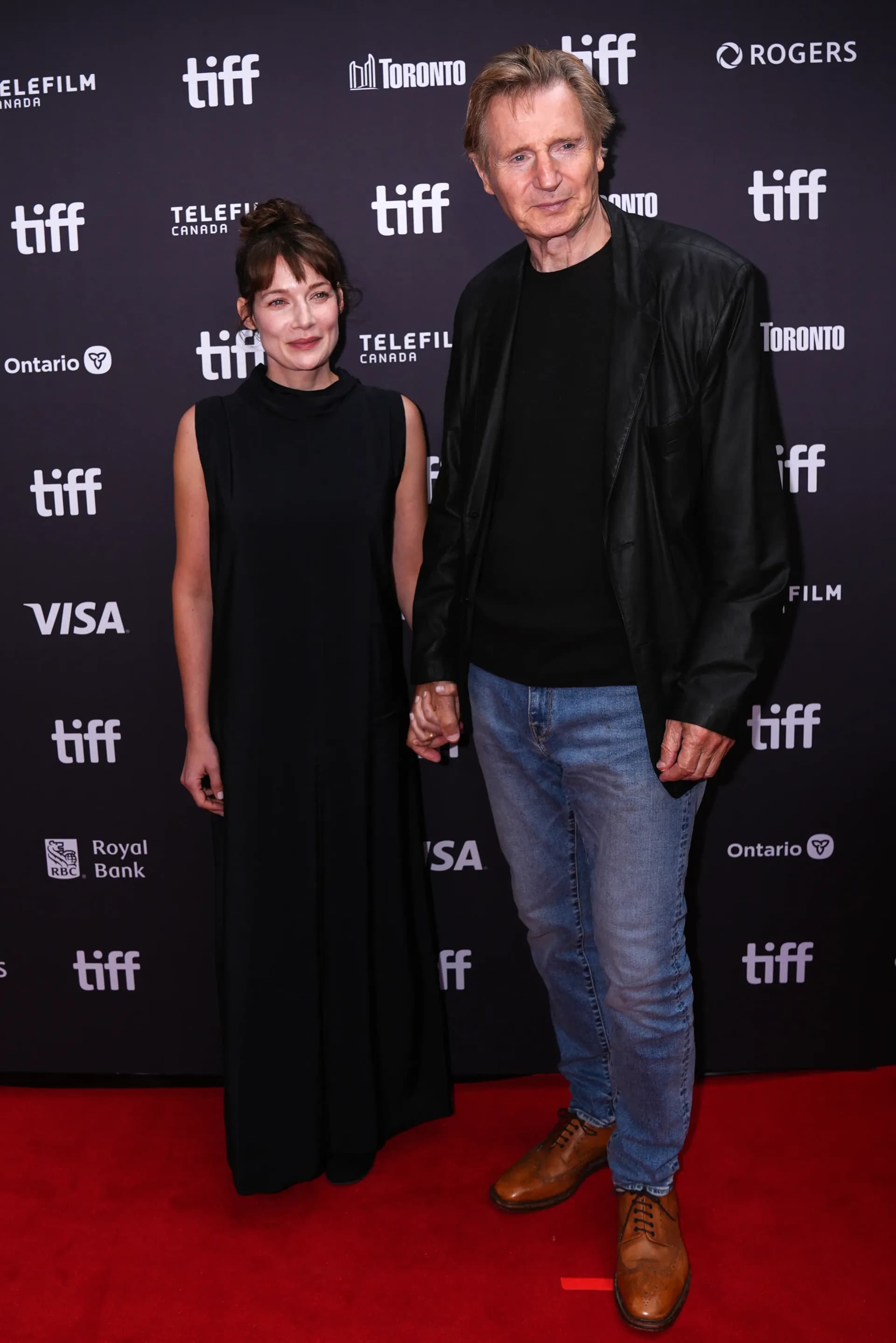 Liam Neeson in a black blazer holding hands with Stella Stocker in a black dress on the Toronto International Film Festival red carpet