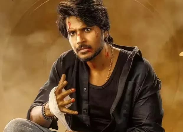 Sundeep Kishan in a dramatic pose, mid-sentence, eyes locked on camera, standing in front of a cinematic backdrop of a heist scene
