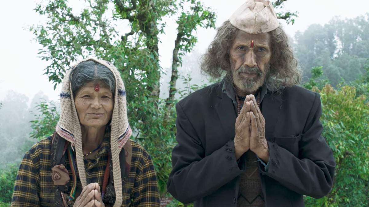 An elderly couple sitting on a wooden bench outside a small house in the hills, looking at the distance. The sky is overcast. The setting is rural Uttarakhand.
