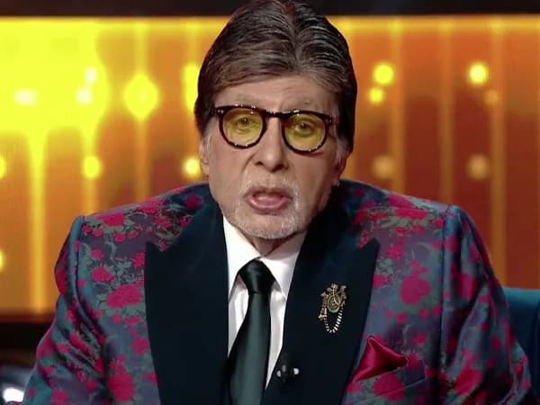 Amitabh Bachchan sitting at the centre of the KBC stage, speaking into a microphone, looking calm and composed.