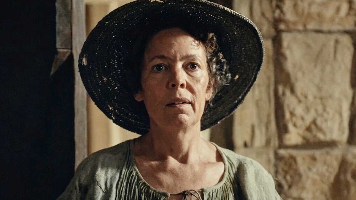 A still from 'Wicker' showing Olivia Colman holding a wicker figure, glowing faintly in dim light.
