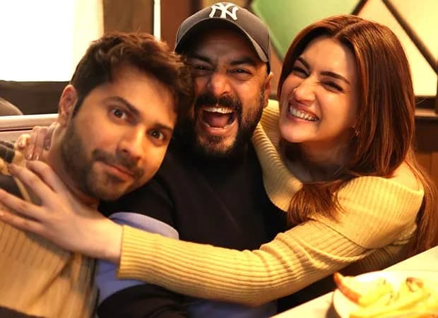 Kriti Sanon in a studio setting, speaking into a microphone, with Varun Dhawan and Amar Kaushik visible in the background