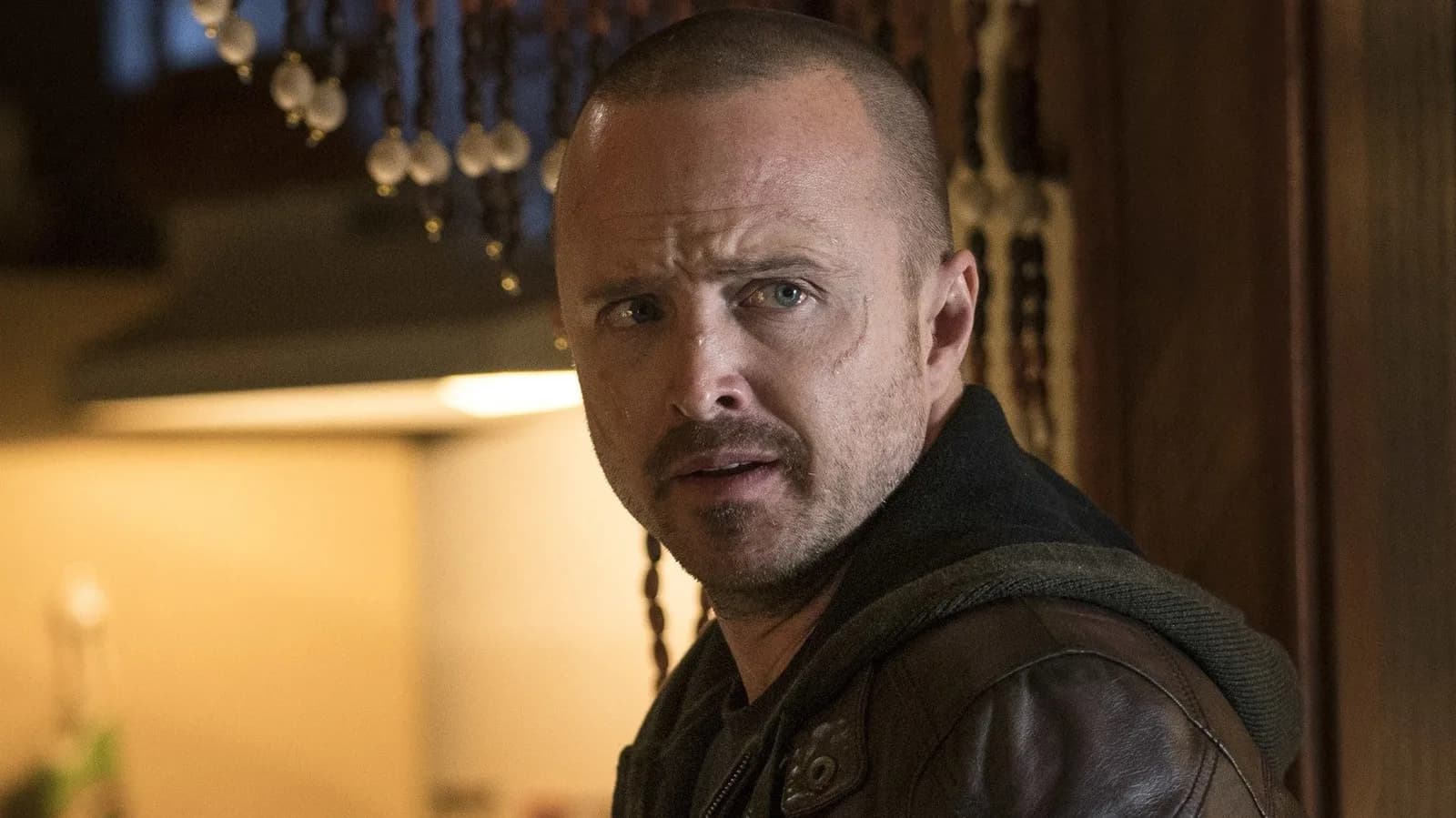 Aaron Paul in a dark suit, face half-melted, standing in a dimly lit club. A blurred reflection of Tom Rhys Harries behind him.
