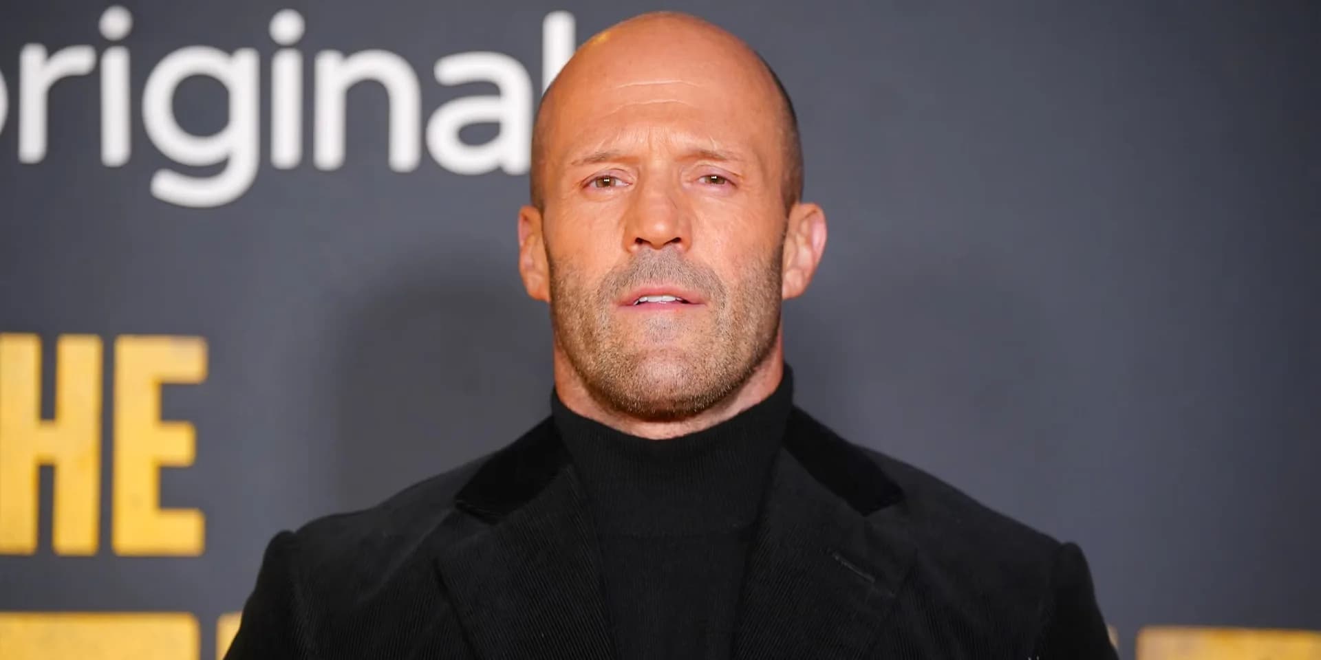 Jason Statham in a dark coat, standing in a dimly lit room, eyes fixed ahead. A single light cuts through the shadows.