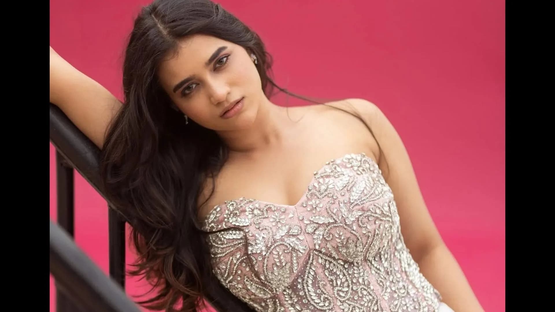 Varupula Madhuri posing in a strapless, heavily jeweled blouse against a bright pink backdrop, hair draped over shoulder