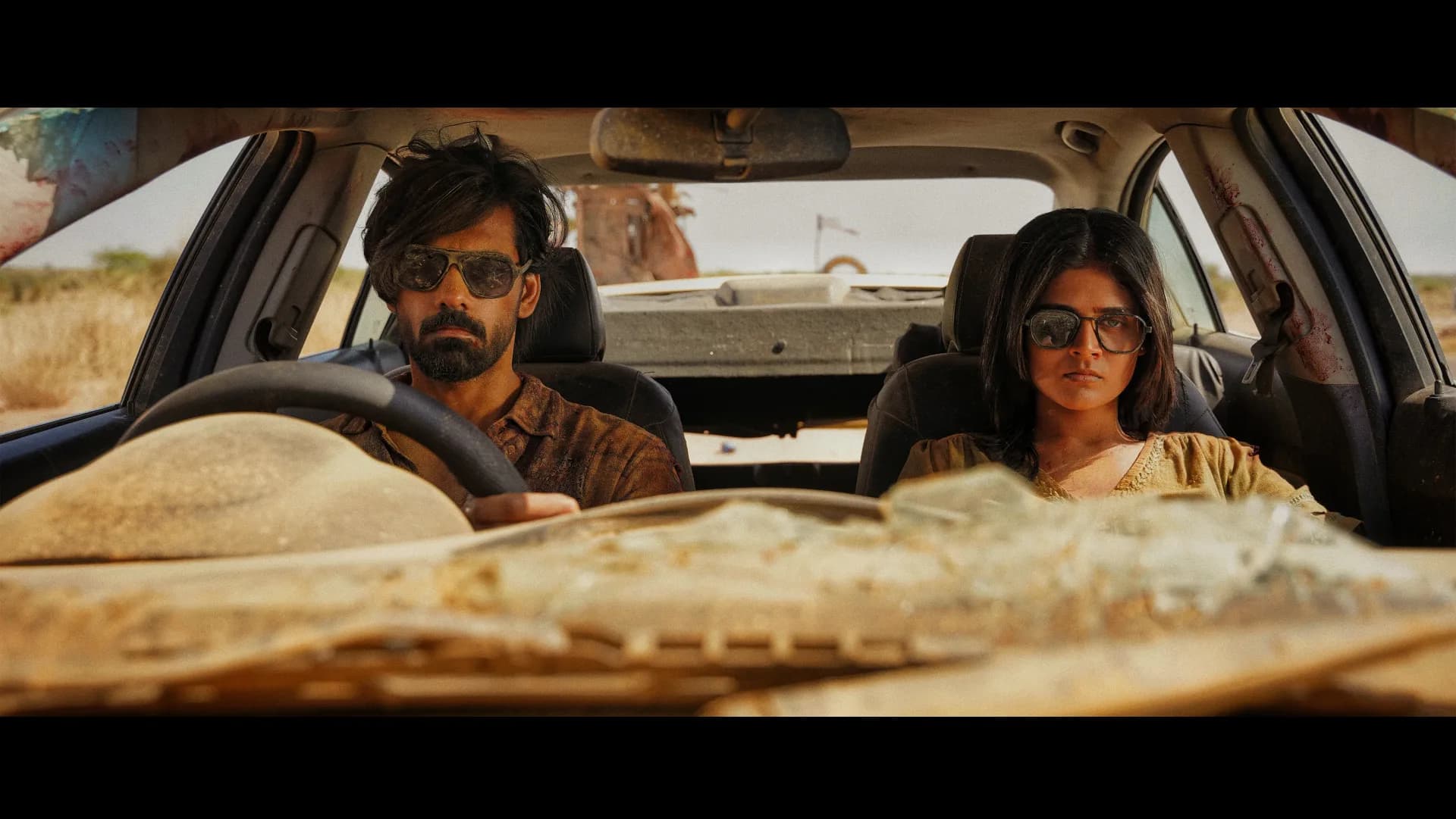 Two vampire figures in sunglasses and dark clothing standing in a desert wasteland under a blazing sun, one holding a knife, the other staring ahead.