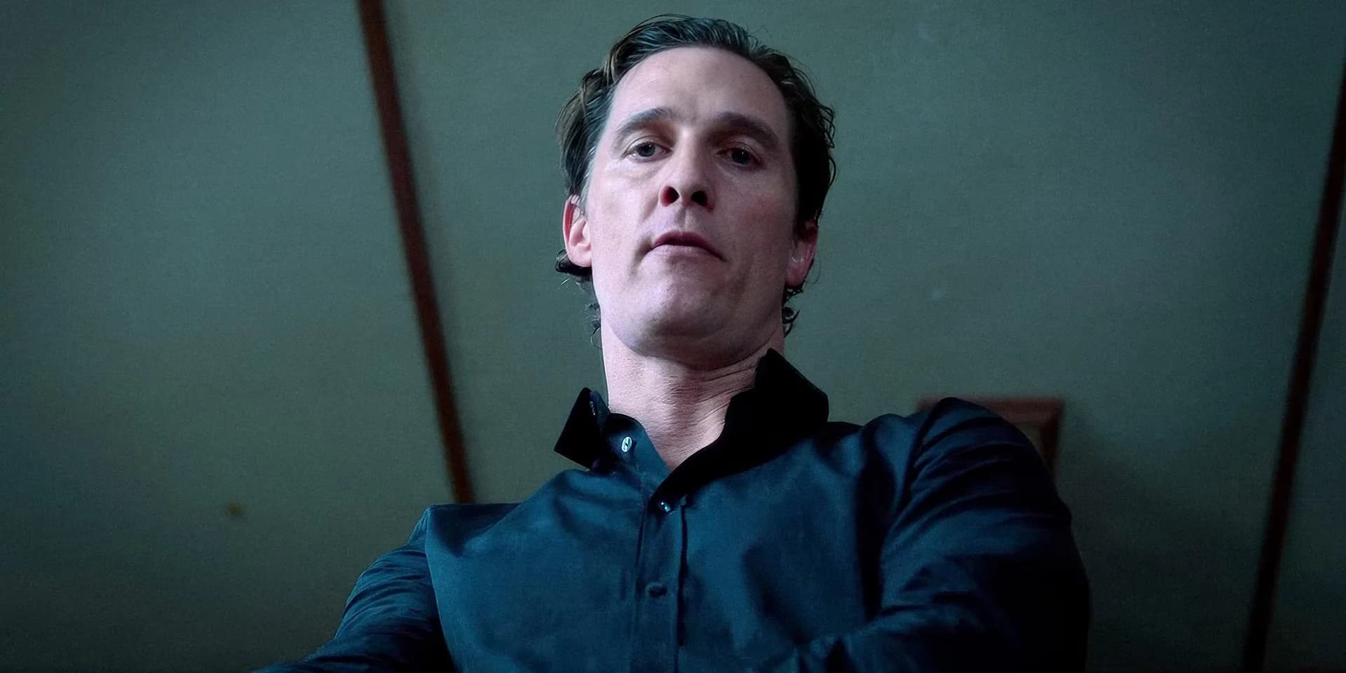 Matthew McConaughey in a dark suit, standing in a dimly lit room, staring at the camera with a calm expression.