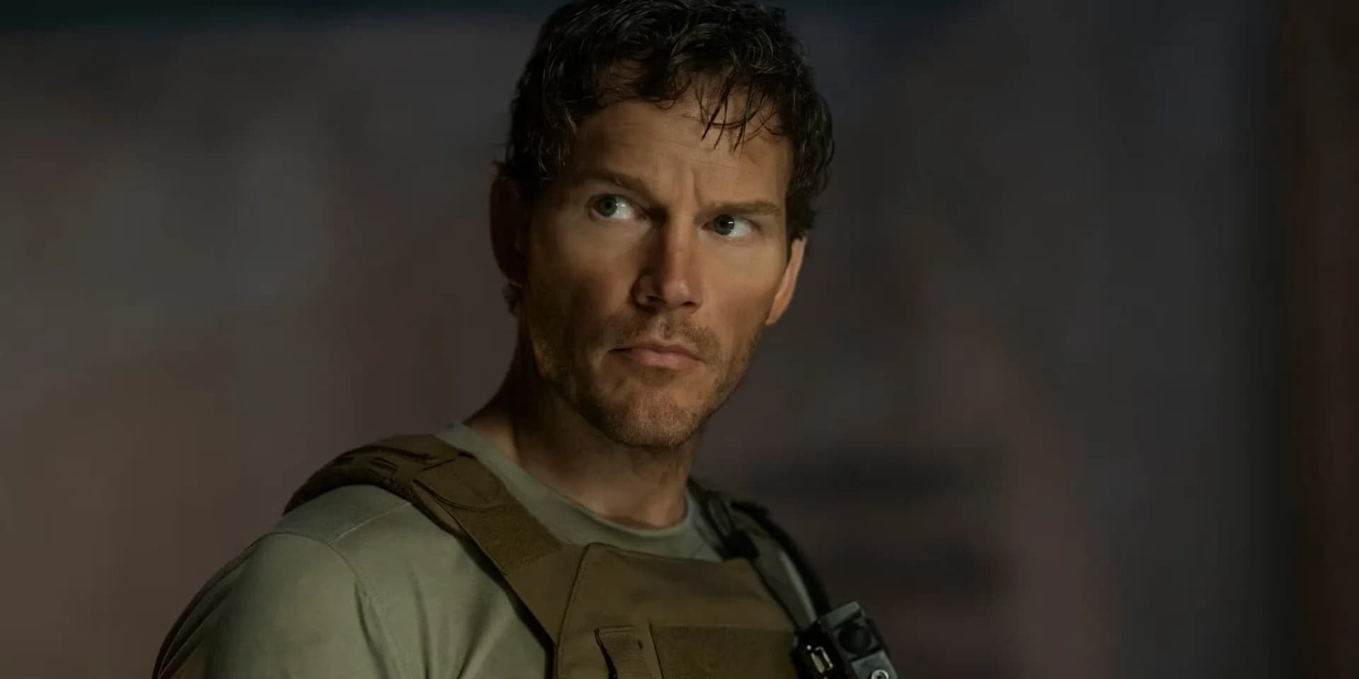 Chris Pratt in a tactical vest, standing in front of a war-torn building, eyes locked ahead.