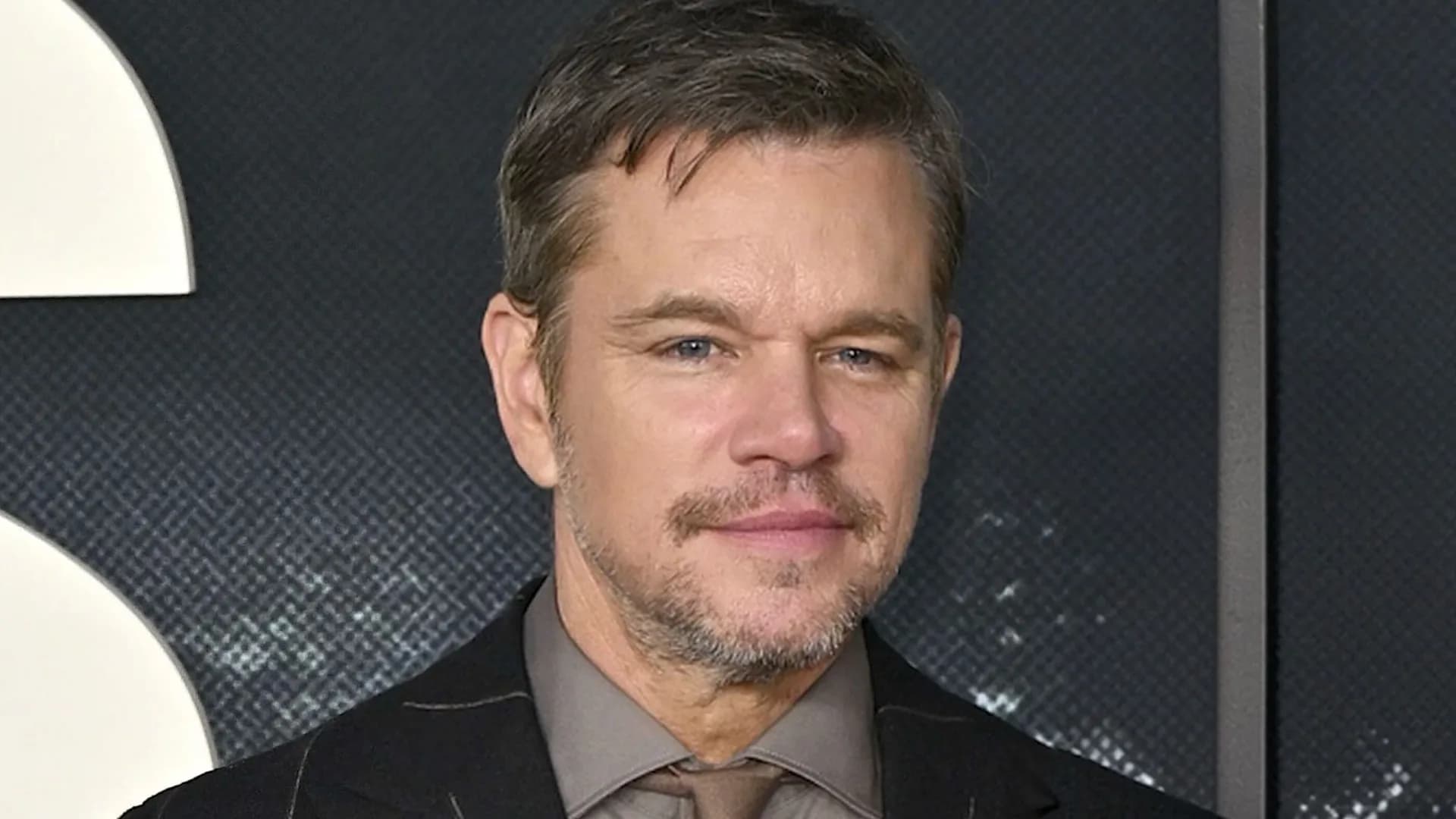 Matt Damon in a suit, standing in front of a city skyline, looking intense.