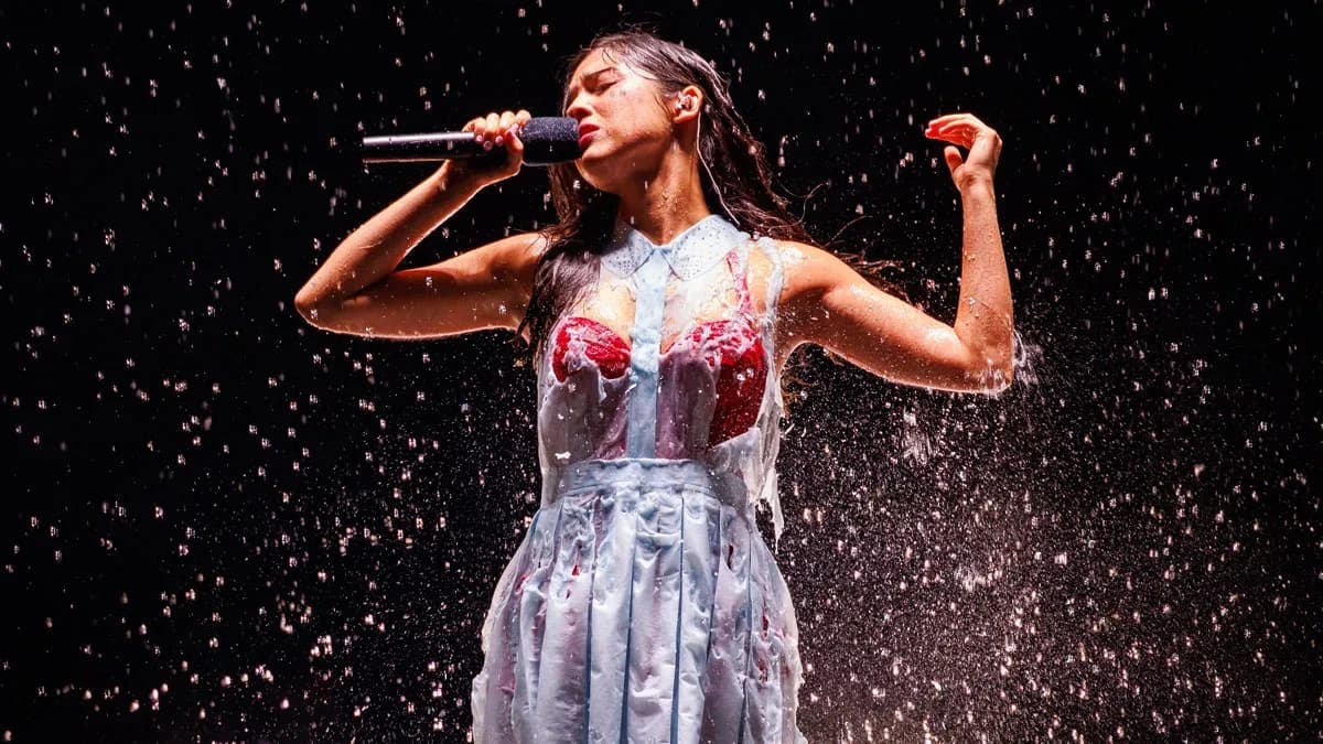 Olivia Rodrigo performing on a B-stage in the rain as her outer dress dissolves to reveal a red lace corseted dress underneath