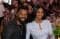 Ciara and Russell Wilson pose together outdoors near a clothesline, both flashing five fingers, with a baby onesie visible among hanging white t-shirts.