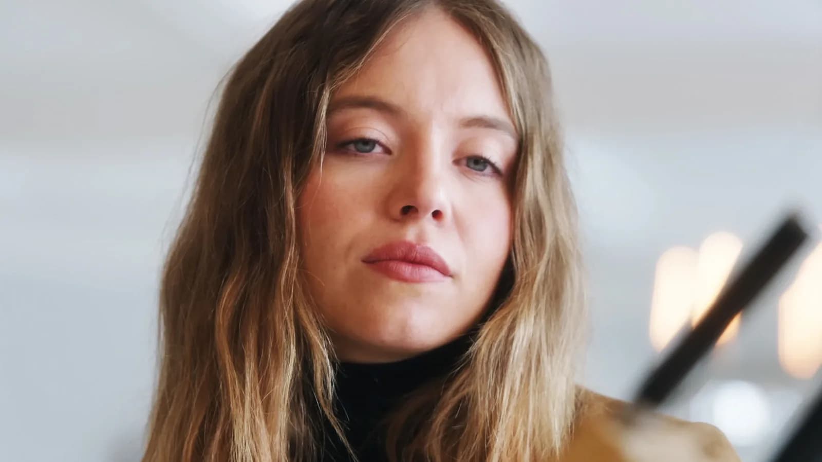 Sydney Sweeney in a tense scene from The Housemaid, dressed in a maid’s uniform, eyes sharp, standing in a dimly lit hallway.