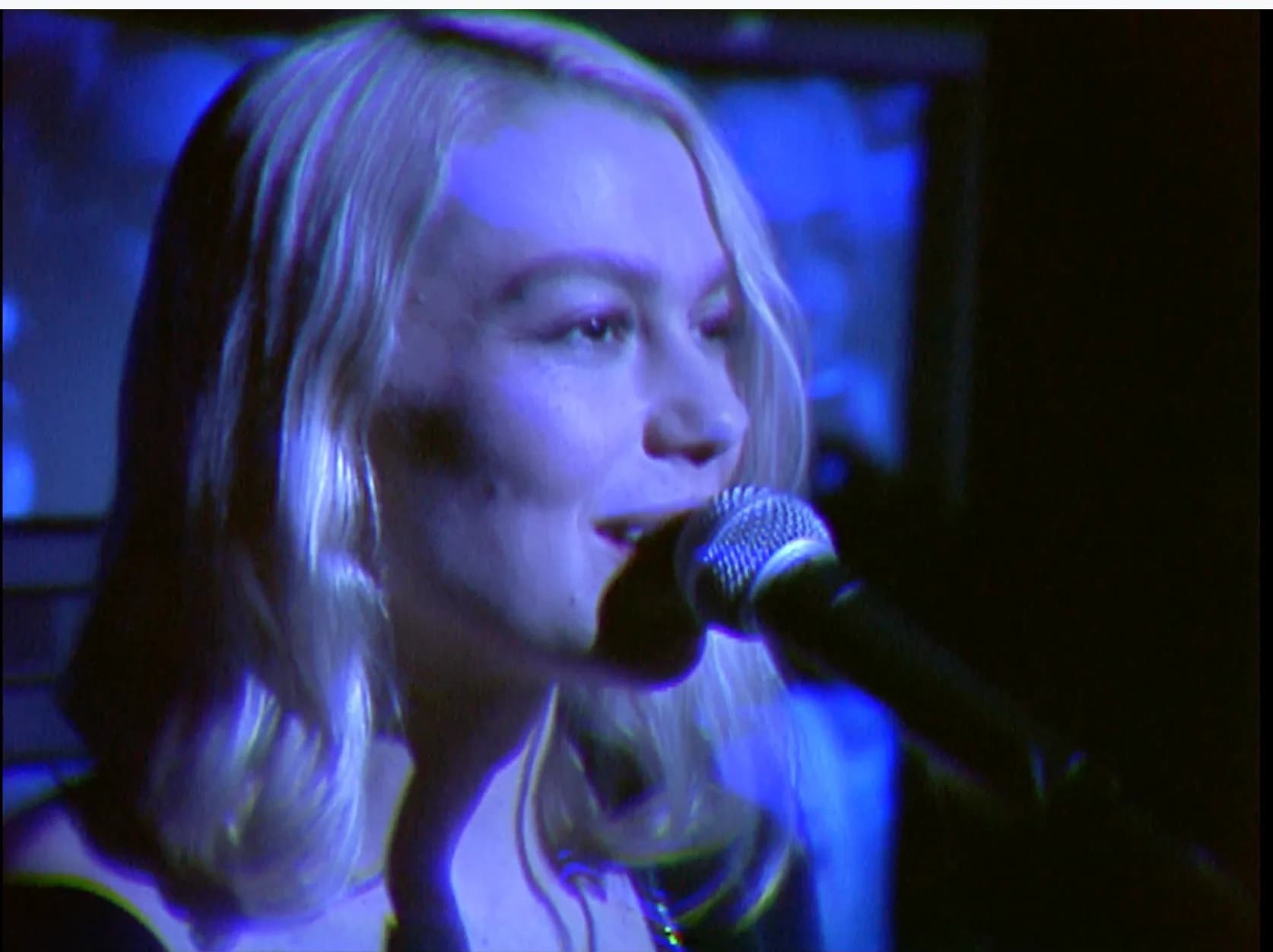 Phoebe Bridgers, Julien Baker, and Lucy Dacus performing together in a dimly lit room with old televisions glowing in the background.