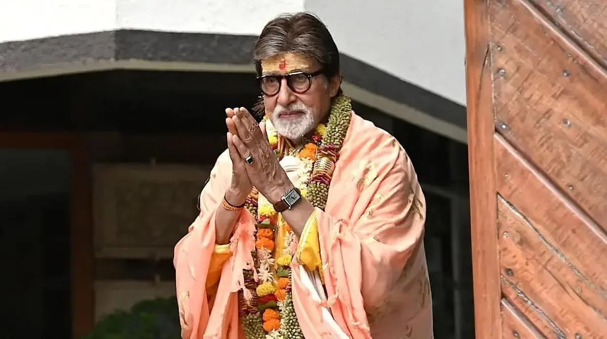 Amitabh Bachchan in a black suit, standing in front of a red curtain, looking directly into the camera with a calm expression.