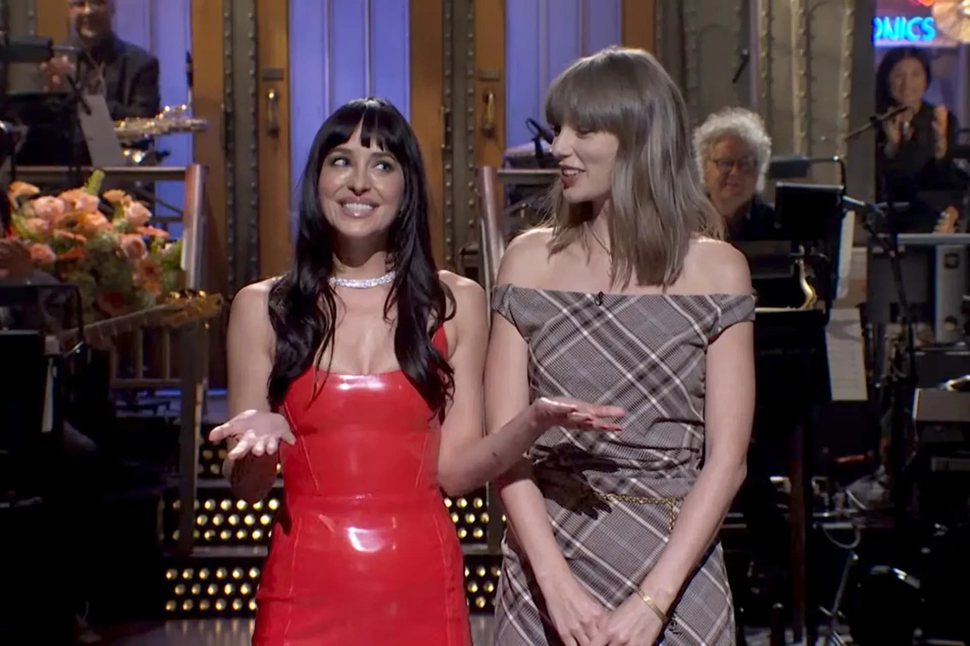 Taylor Swift in a plaid off-the-shoulder mini dress, standing next to Dakota Johnson on the SNL stage.