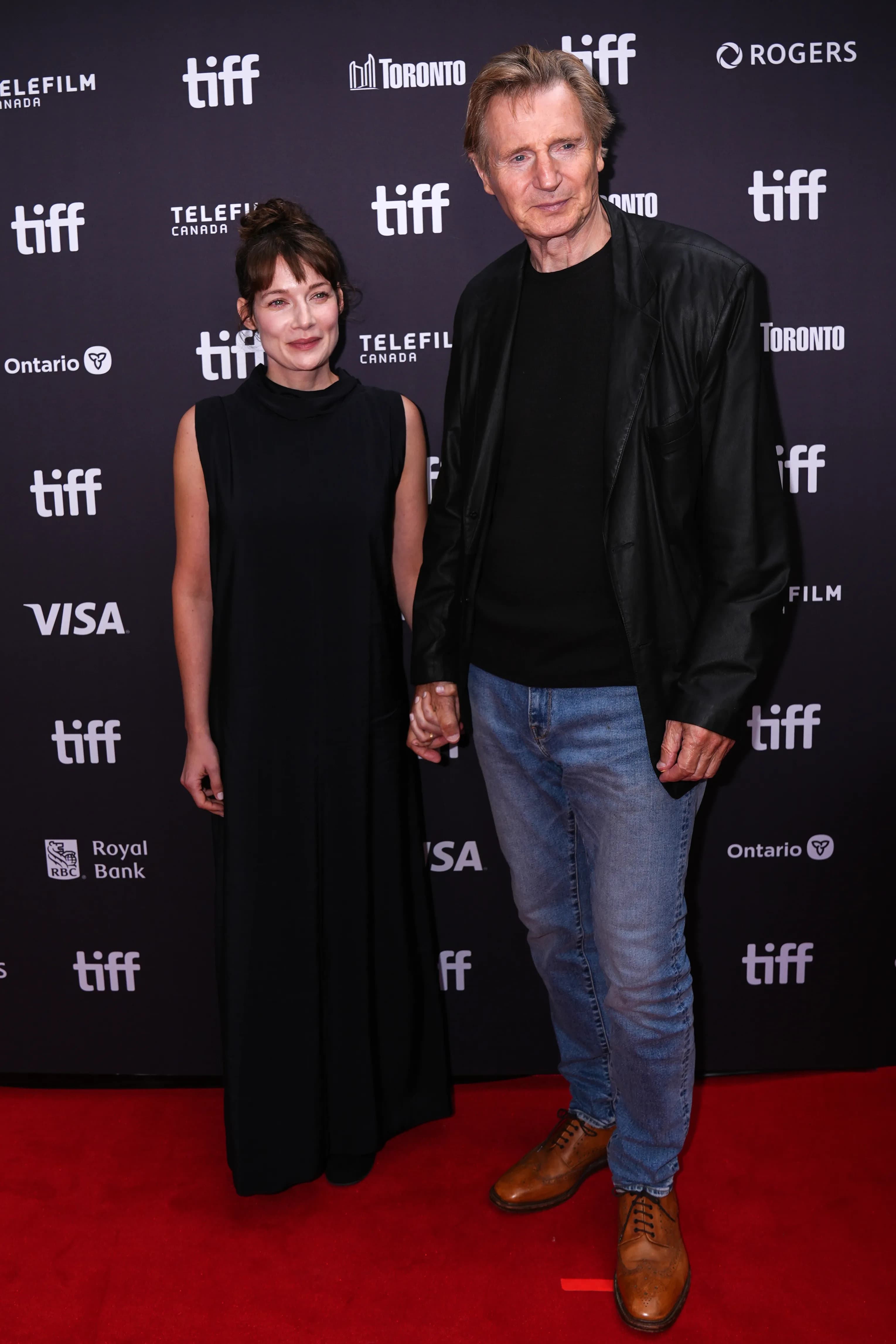 Liam Neeson in a black blazer holding hands with Stella Stocker in a black dress on the Toronto International Film Festival red carpet