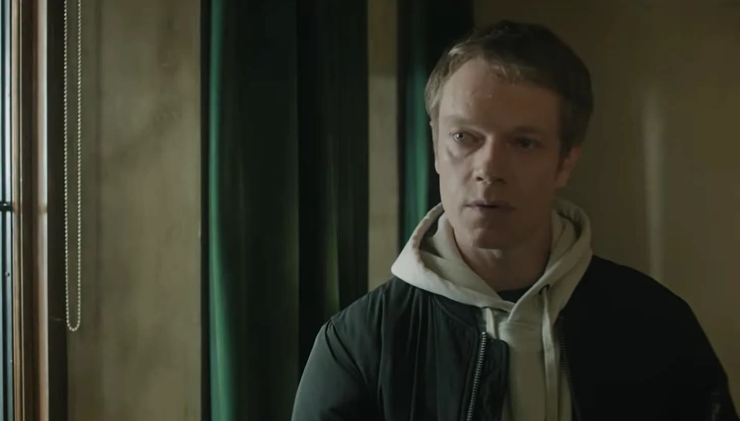 Alfie Allen as Nolan Reeves in 'The Institute' Season 2, standing in a dimly lit room, expression unreadable.