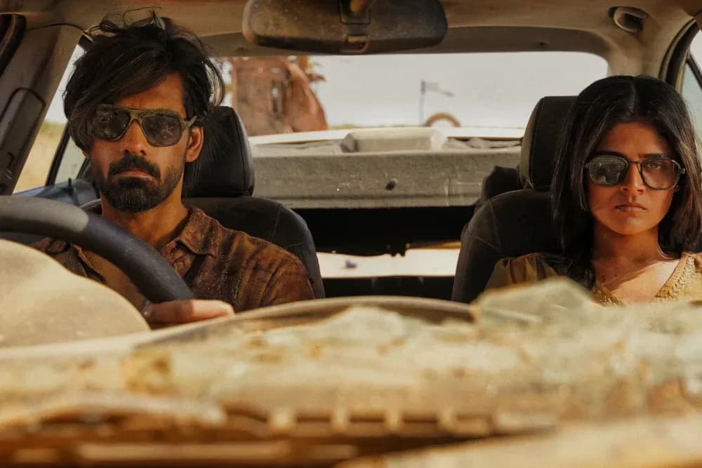 A still from 'Half' showing two figures in silhouette, backlit by a dusty sun, mid-fight. Blood sprays across cracked earth.