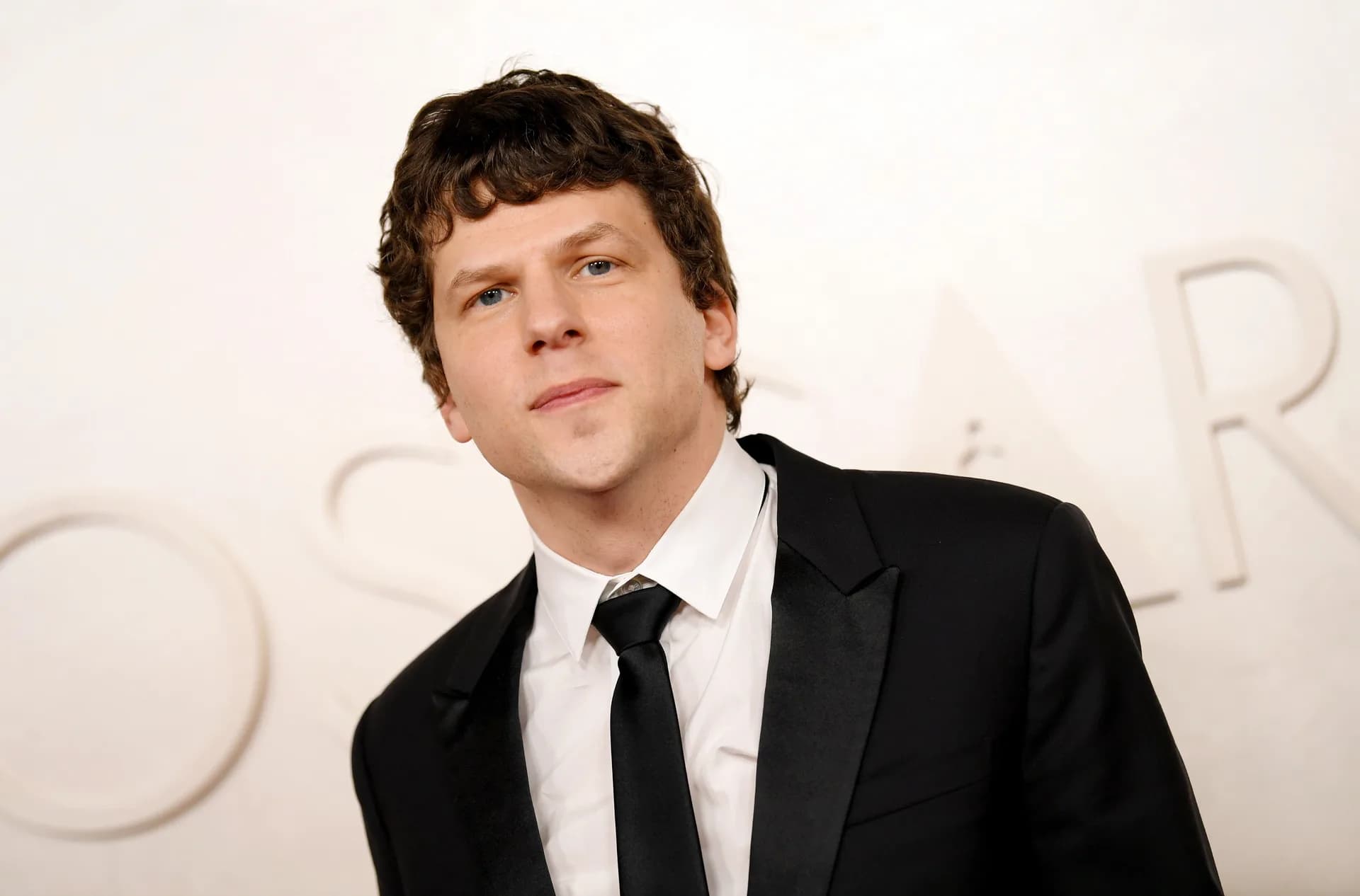 Jesse Eisenberg in a black turtleneck, standing in front of a grey wall, looking thoughtful.