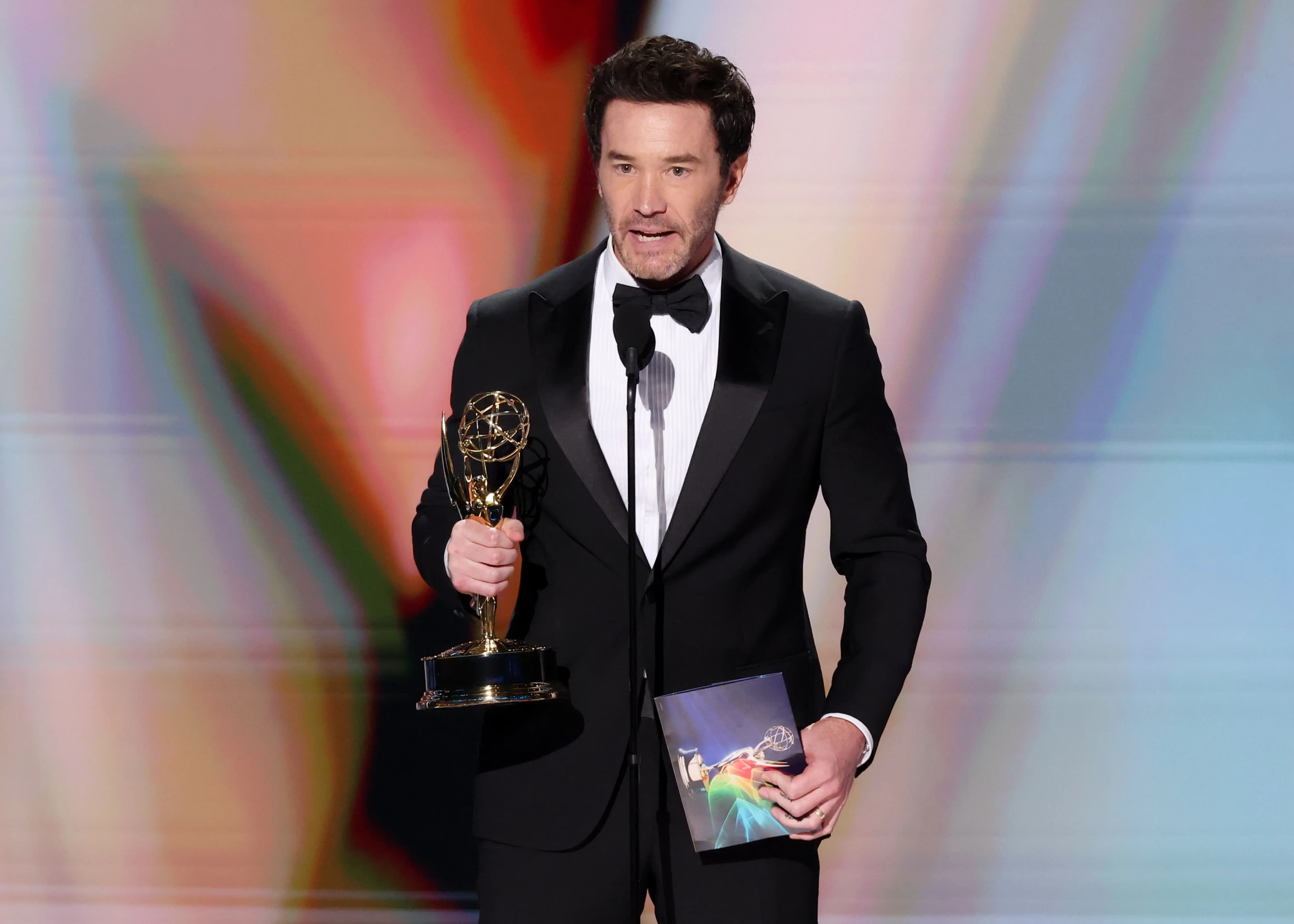 Tom Pelphrey holding his Emmy Award statuette on stage at the Peacock Theater.
