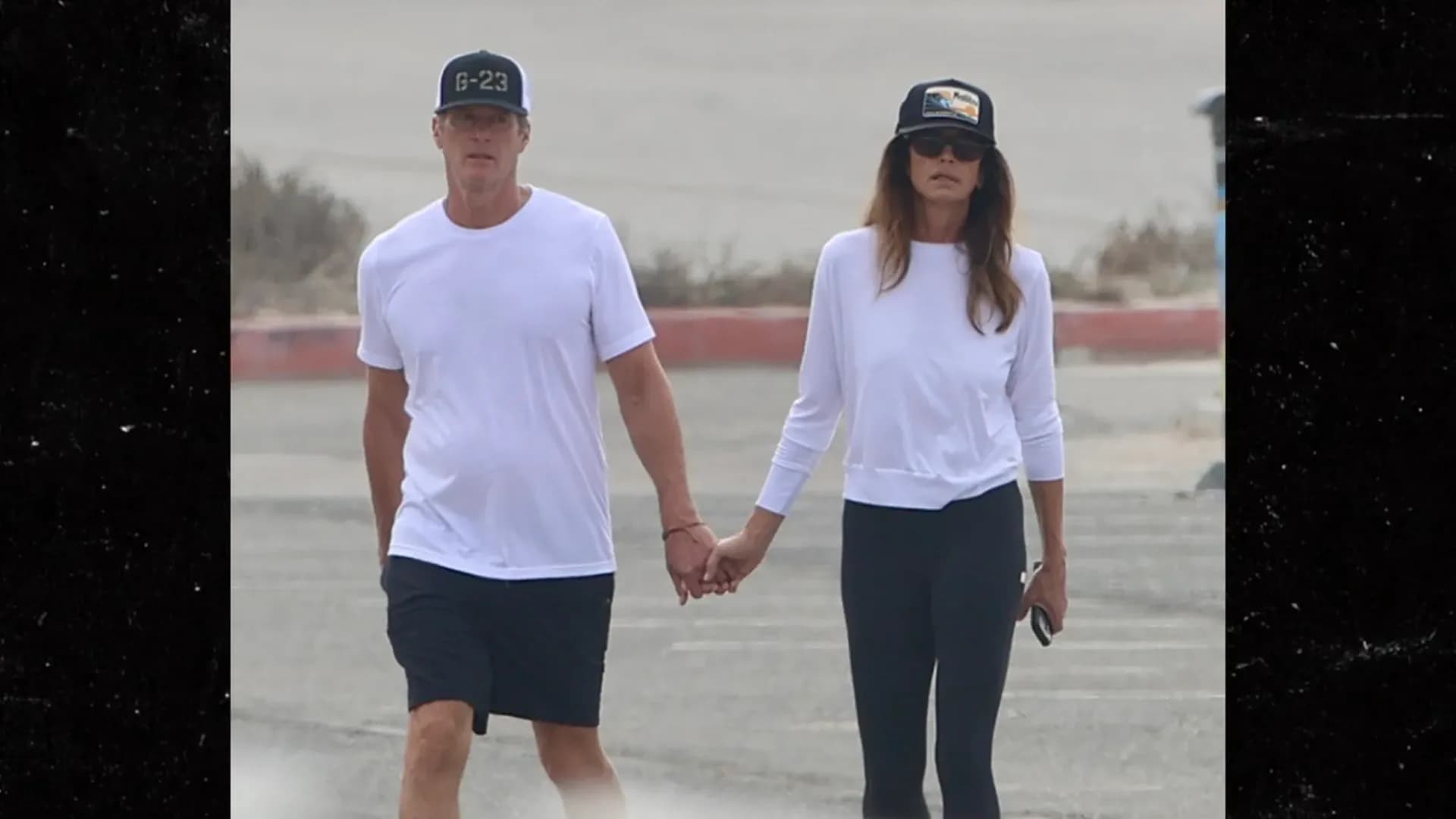 Cindy Crawford and Rande Gerber walking hand-in-hand on Zuma Beach, wearing white shirts and caps, silhouetted against the ocean.