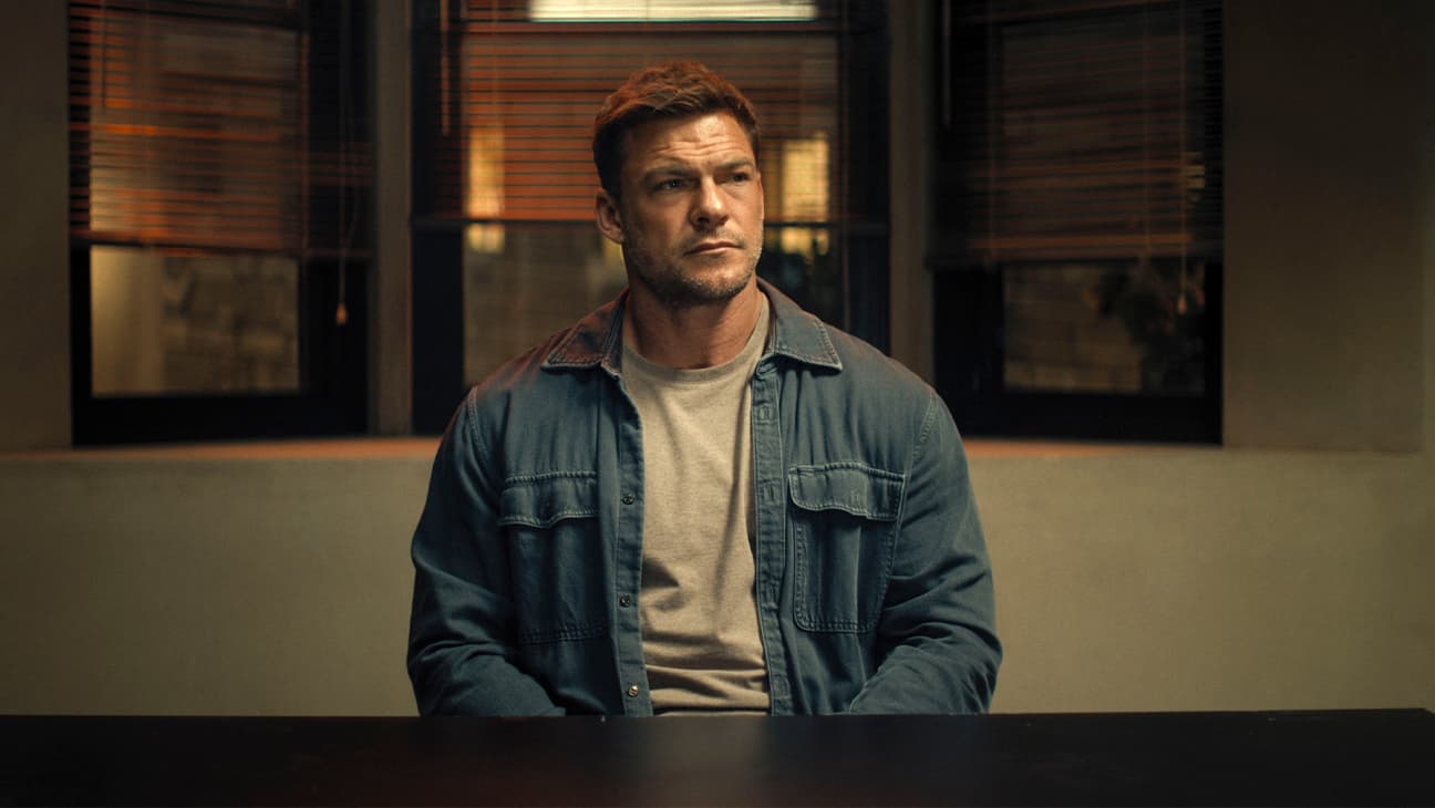 Alan Ritchson speaking at a microphone with a Reacher poster in the background