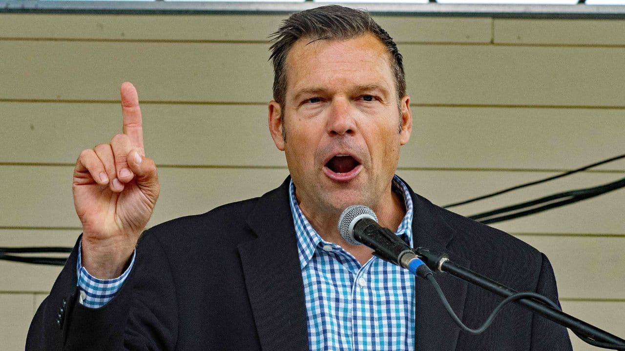 Kansas Attorney General Kris Kobach speaking at a press podium with state flags behind him