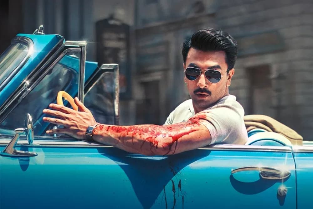 Ranbir Kapoor in a turquoise vintage car, sunglasses, blood streaking from shoulder to wrist, gold watch on injured arm