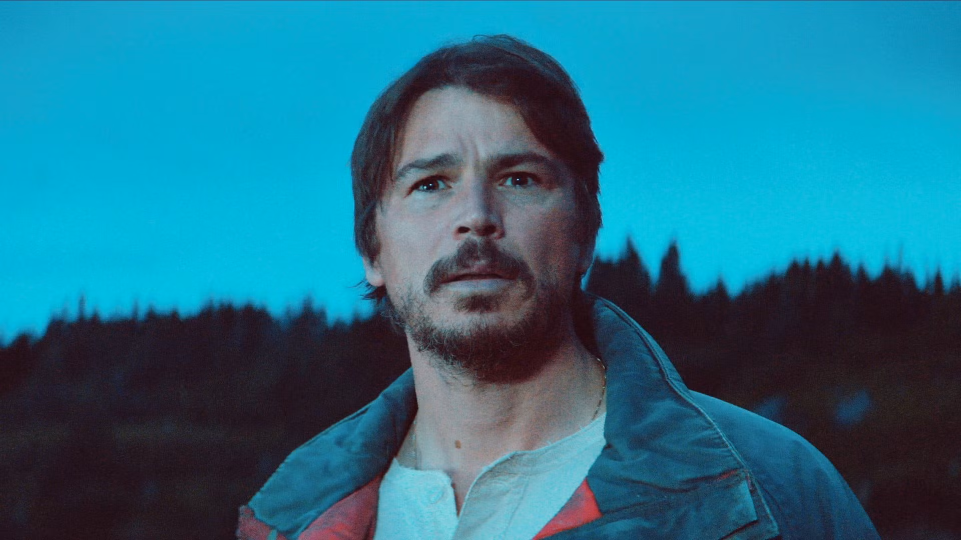 Josh Hartnett in a weathered coat, standing at the edge of a fog-drenched coastline, looking out at dark water.