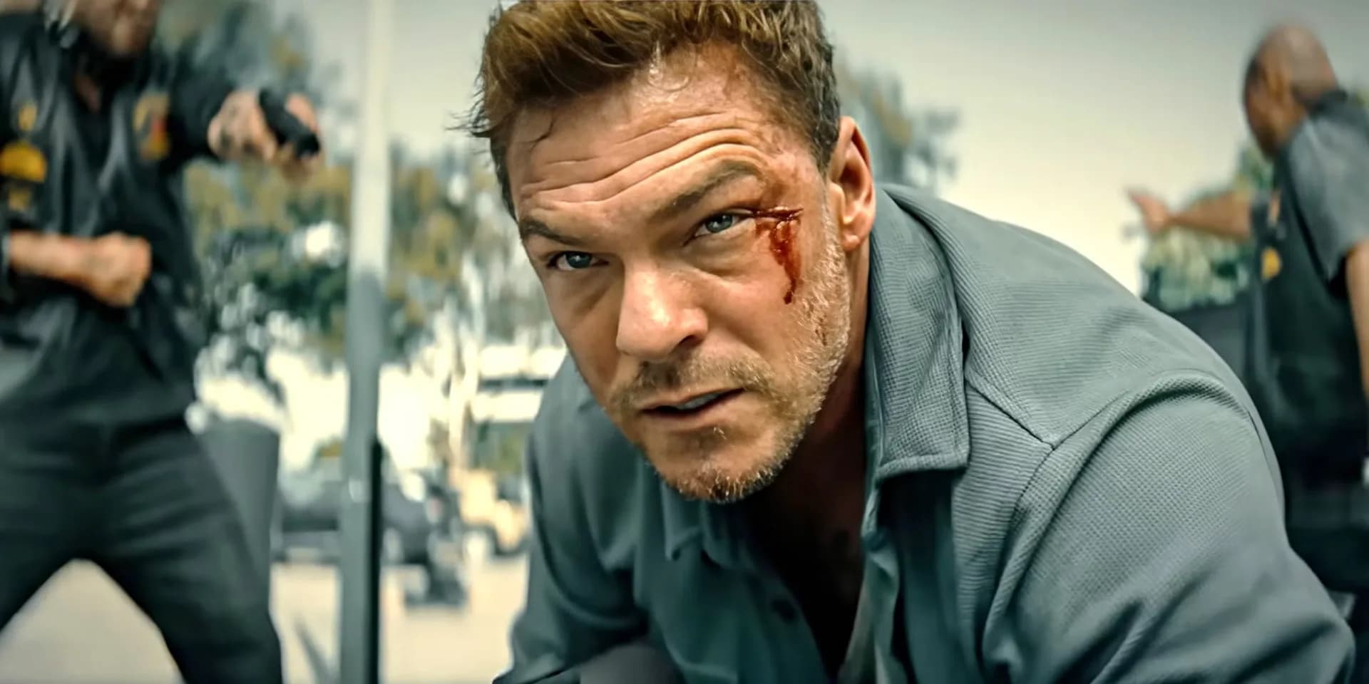 Alan Ritchson in a gritty action pose, holding a weapon, standing in front of a stormy sky.