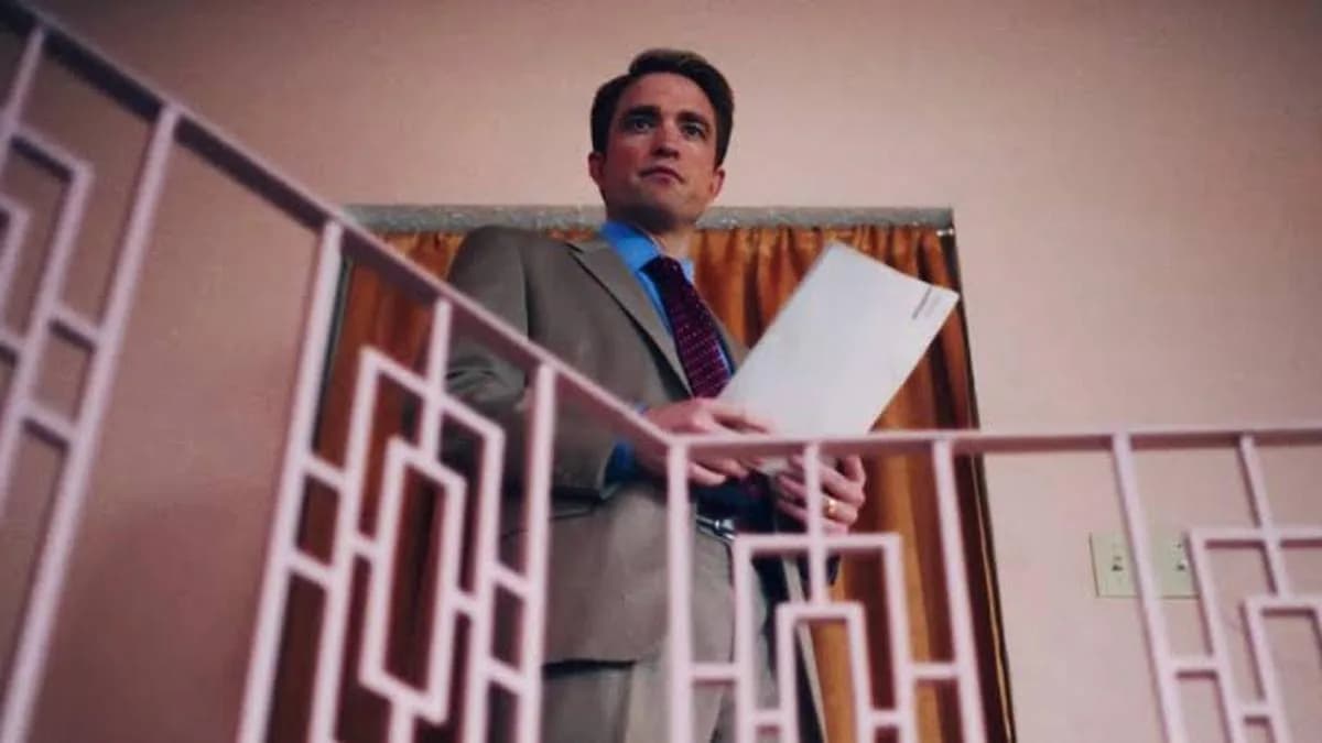 Robert Pattinson in a grey suit, staring into the camera with intense focus, in a dimly lit room. The image has a grainy, vintage TV aesthetic.