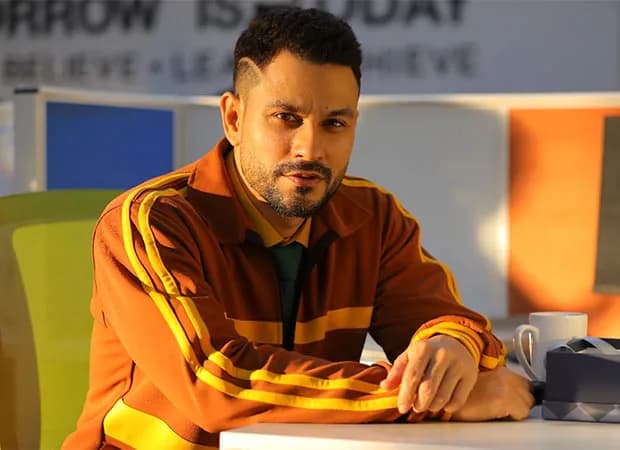 Kunal Kemmu standing on the Vibe set, illuminated by colorful lighting, looking toward the camera.