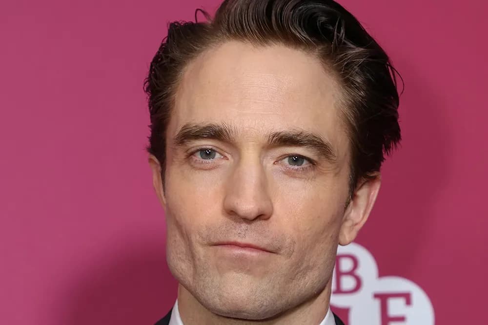 Robert Pattinson at the London Film Festival premiere of Primetime, wearing a dark grey suit and slicked-back hair, smiling at the audience.