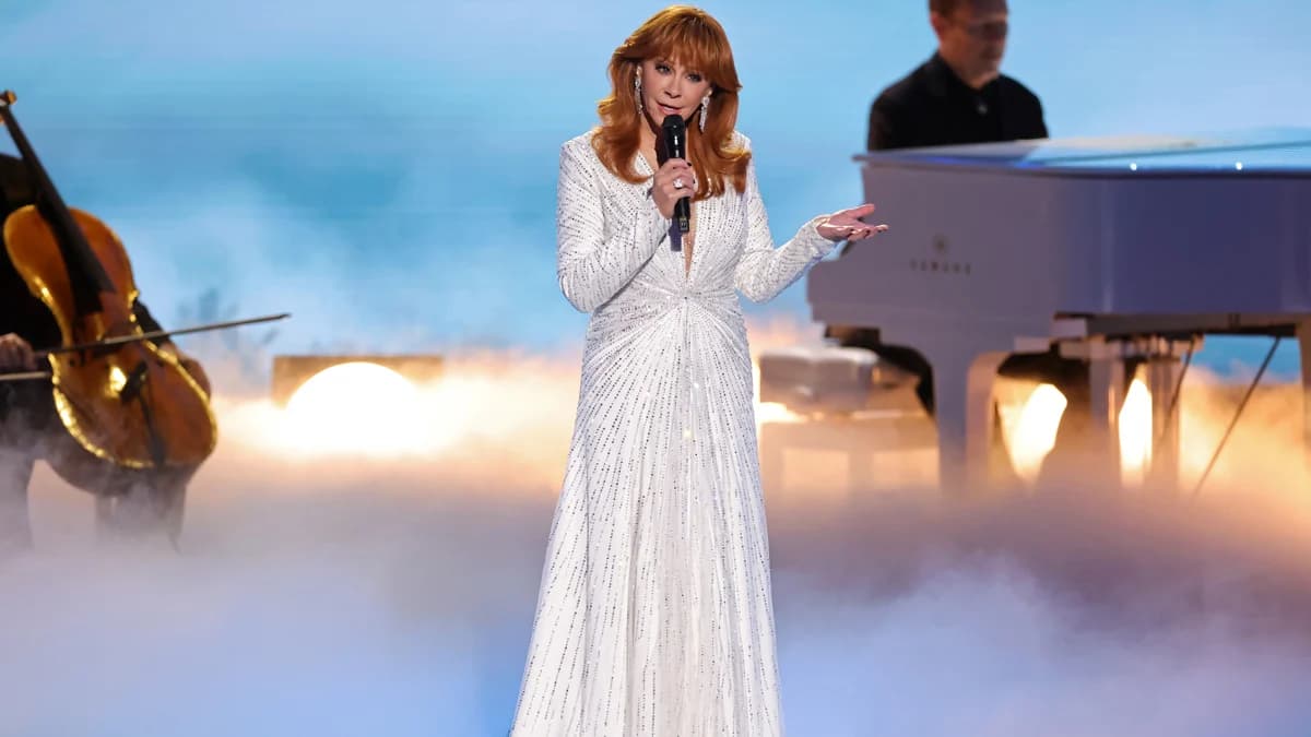 Reba McEntire in a sparkling white gown singing on the Emmy Awards stage with photographs of Dolly Parton projected behind her.