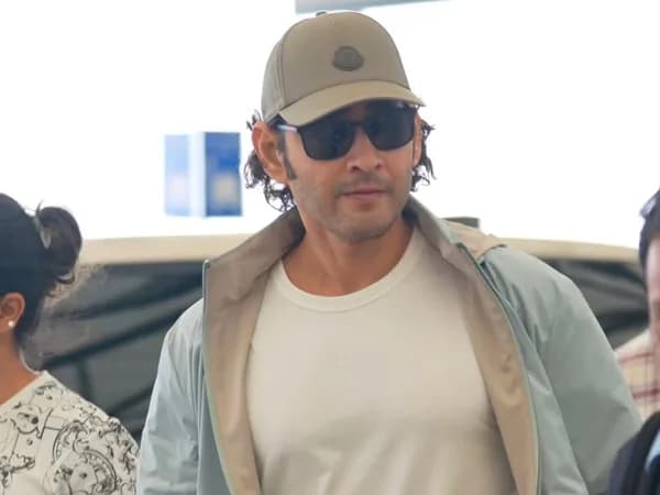 Mahesh Babu at an airport wearing a white T‑shirt, beige cap and sunglasses, showing a clean‑shaven face.
