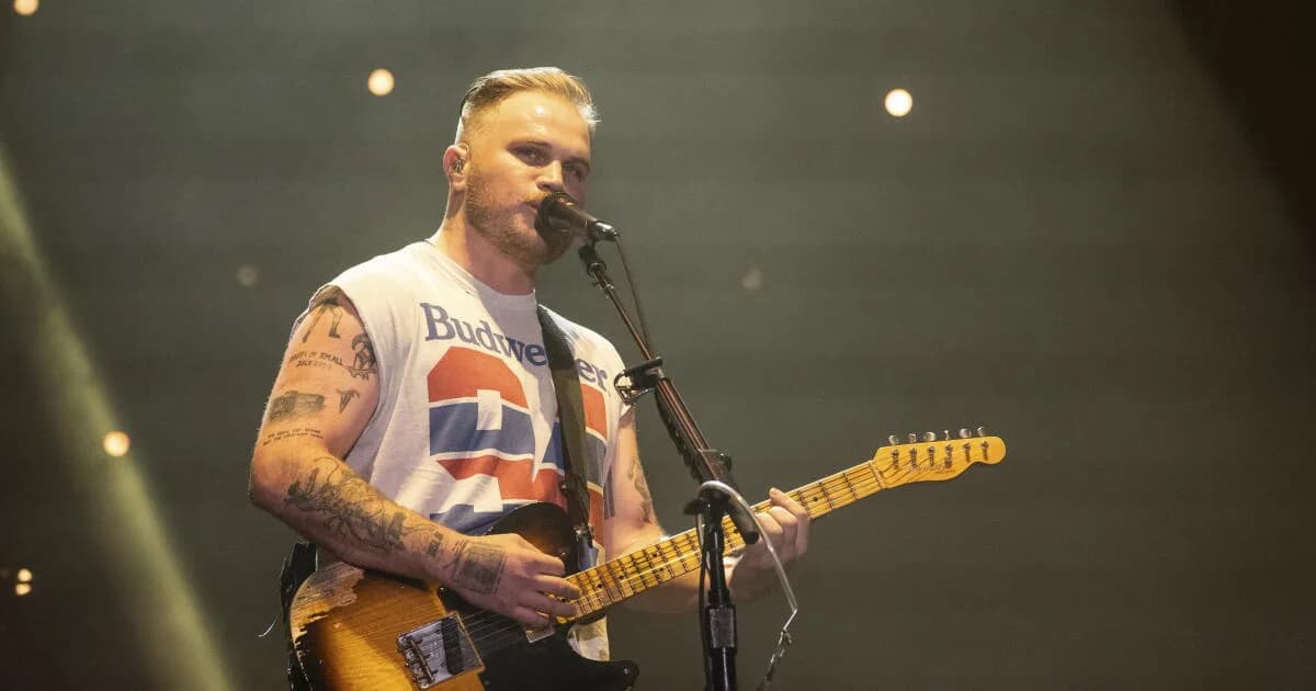 Zach Bryan performing on stage at Gillette Stadium, wearing a black T-shirt with 'Free Palestine' and 'Appeal to Heaven' printed on it.