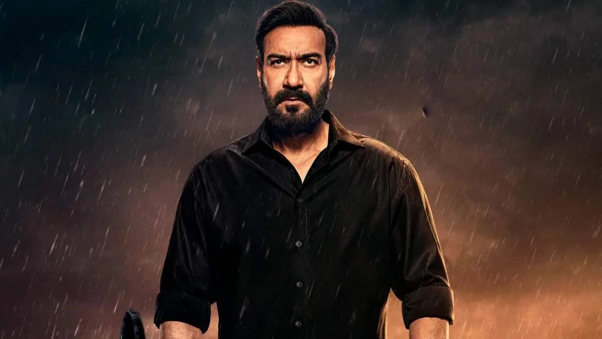 Ajay Devgn in a tense expression, standing against a dark background, holding a phone.