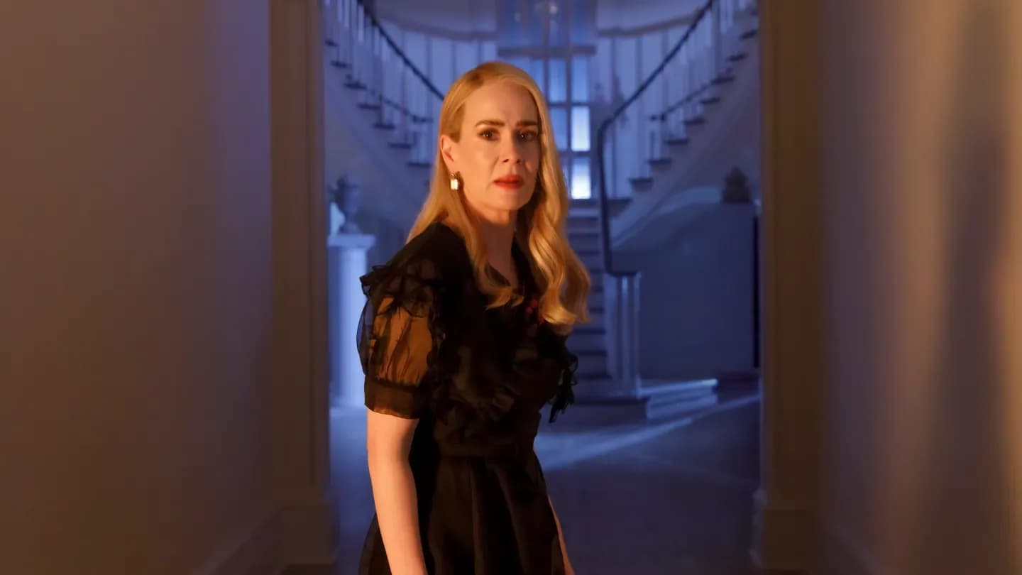 Jessica Lange as Fiona Goode, standing in a dimly lit room, eyes glowing red, holding a staff. Behind her, shadows of other witches loom.