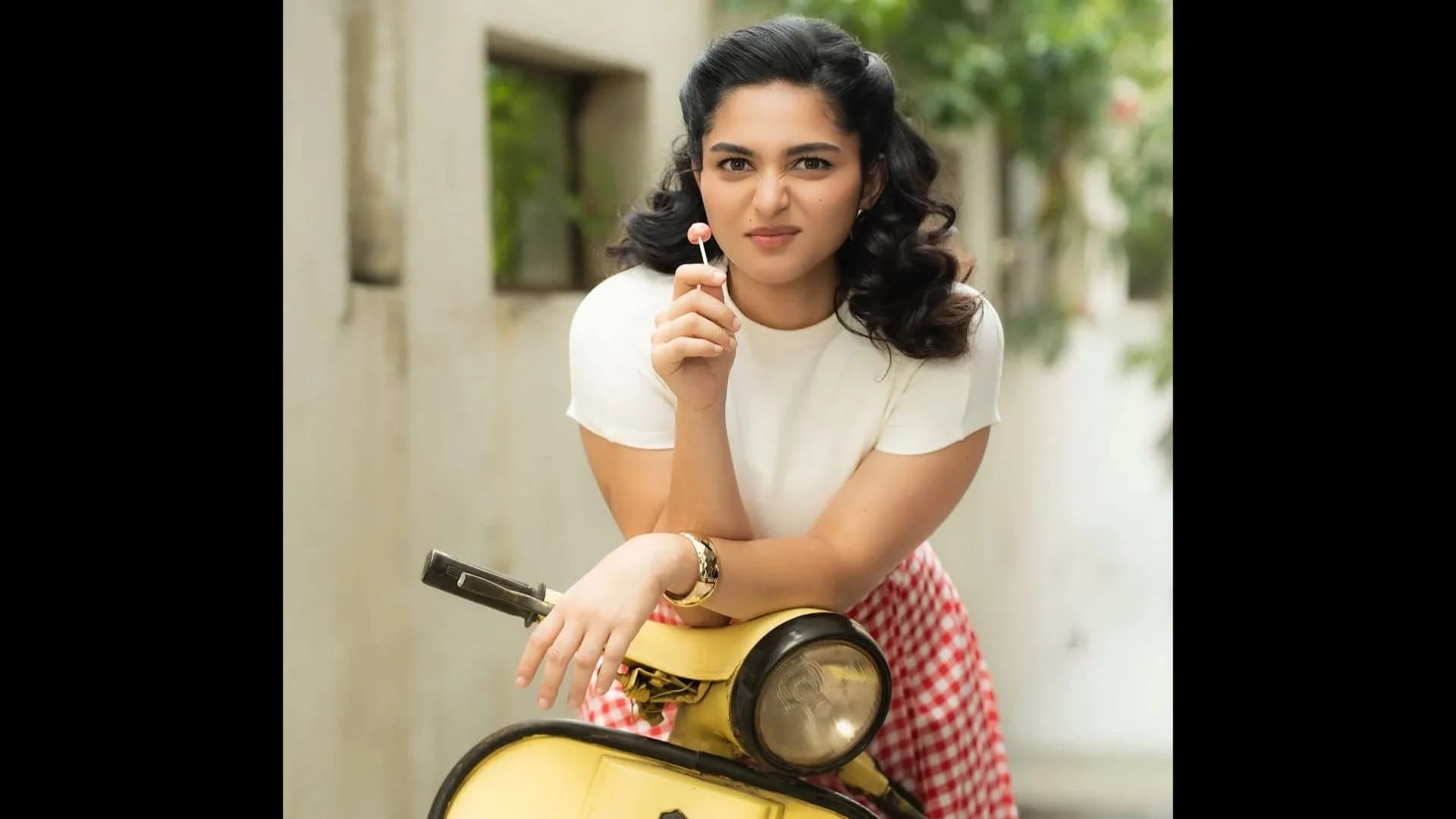 Kayadu Lohar smiling with a lollipop, wearing a white top and pink trousers, posing beside a vintage Bajaj Chetak scooter.