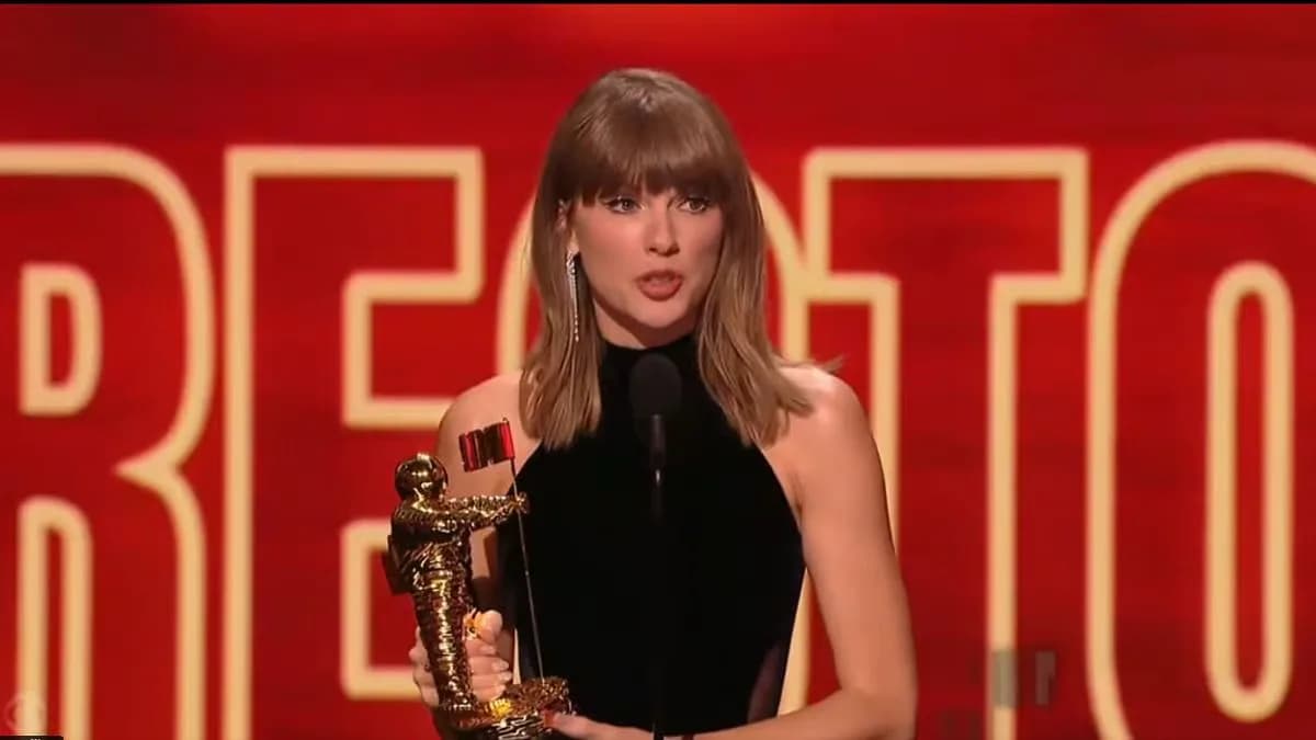 Taylor Swift holding a Moon Person trophy on the VMA stage, with a backdrop of glittering lights.