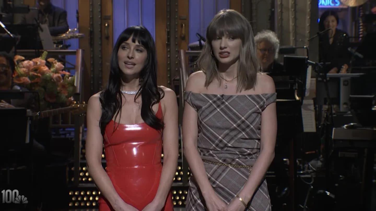 Taylor Swift and Dakota Johnson laughing on stage during an SNL monologue, with a blurred crowd in the background.