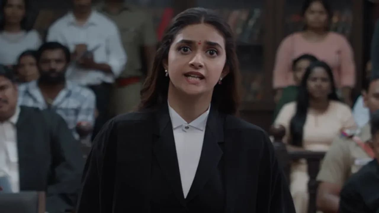 Keerthy Suresh in a courtroom scene, eyes locked on Mysskin, who stands tall in a suit, expression unreadable.