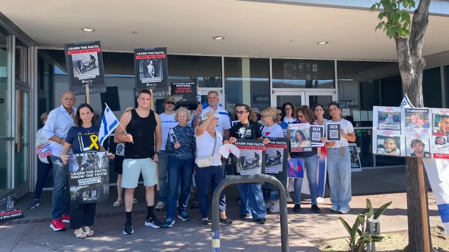 A group of protesters holding signs and flags outside a movie theater, with the film title 'NAZA' visible on a marquee.